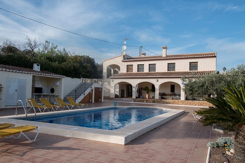 Spaans huis met muren in gebroken wit, een veranda met bogen in hetzelfde gebroken wit, oranje dakpannen en op de voorgrond een zwembad met daarnaast vier ligstoelen.