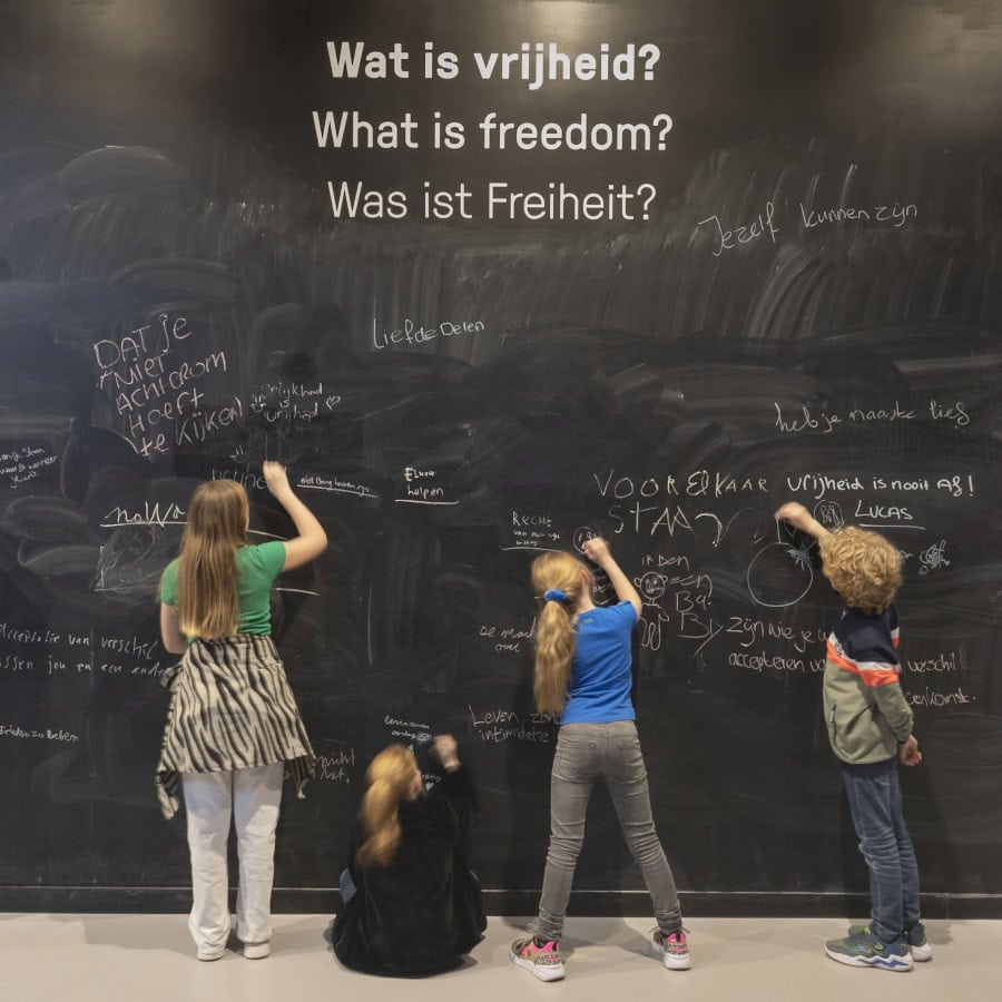 Vier kinderen krijten op een zwarte muur, waar bovenaan "Wat is vrijheid?" en daaronder de Engelse en Duitse vertalingen in witte letters op staat. De kinderen zijn op hun rug zichtbaar, drie kinderen staan te krijgen, en eentje zit op de grond. 