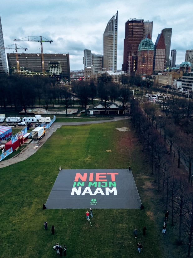 Grasveld van het Malieveld met links achter enkele caravans en een deel van een circustent. Op de voorgrond is de banner met 'Niet in mijn naam' te zien, met enkele mensen erbij. Op de achtergrond de hoogbouw die kenmerkend is voor het centrum van Den Haag.